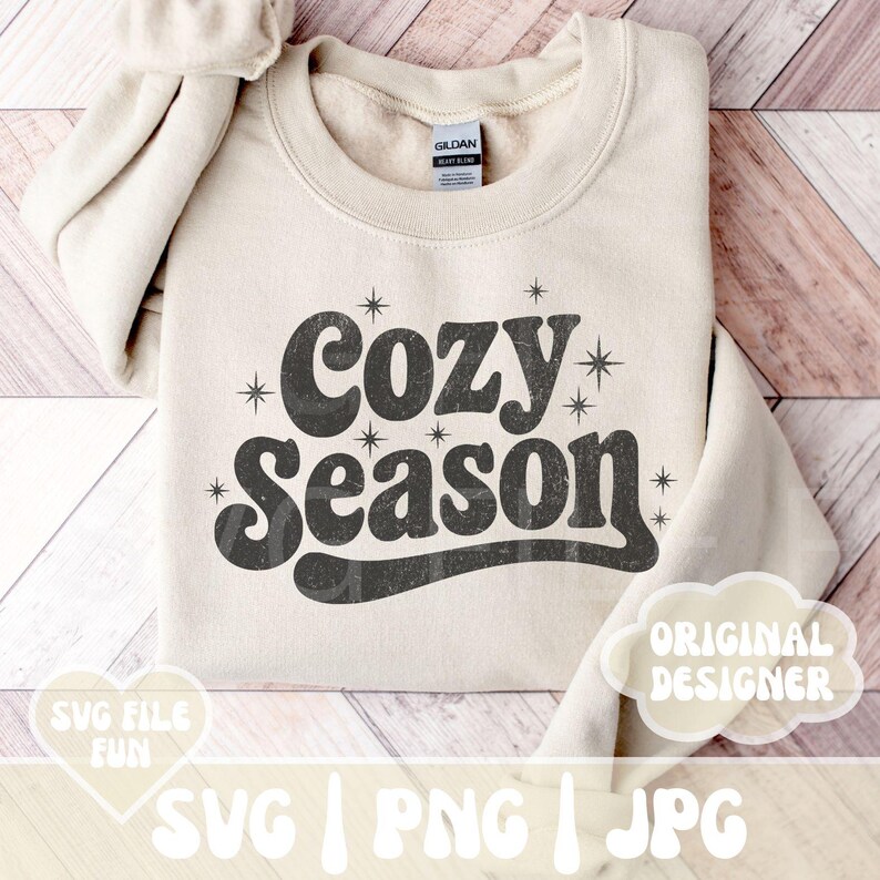 Puede incluir: Sudadera beige con la frase "Cozy Season" en una fuente retro. El texto es de color gris oscuro con detalles de estrellas. La sudadera est&aacute; doblada sobre una superficie de madera clara.