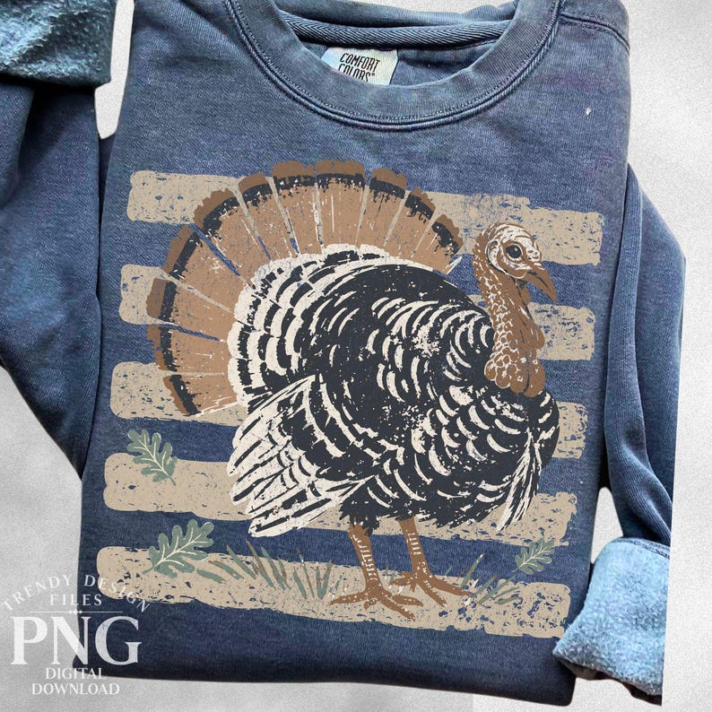 May include: A blue long-sleeve sweatshirt featuring a graphic of a turkey with brown and black feathers. The design includes horizontal beige stripes and small green leaf accents. The inner neck label displays the text Comfort Colors.
