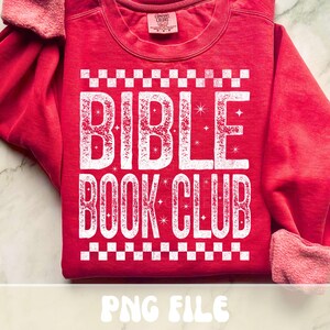 以下が含まれることがあります： 白い文字で「BIBLE BOOK CLUB」と書かれた赤いスウェットシャツ。テキストの上と下に市松模様があります。スウェットシャツはクルーネックと長袖です。