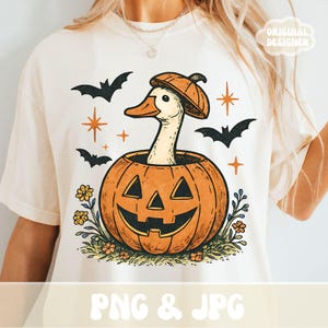 May include: Cream-colored t-shirt featuring a Halloween-themed graphic. The design includes an orange pumpkin with a smiling face, a goose wearing a hat, bats, and floral accents. The text "PNG & JPG" is at the bottom.