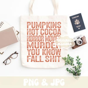 Puede incluir: Bolso tote crema con el texto naranja "PUMPKINS HOT COCOA HORROR MOVIES MURDER YOU KNOW FALL SHIT". Sobre una superficie blanca con pasaporte, c&aacute;mara, gafas y vegetaci&oacute;n.