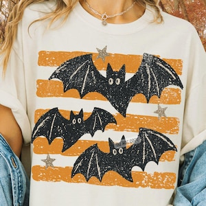 May include: A cream-colored t-shirt featuring a graphic print of three black bats flying across horizontal, distressed orange stripes. The design includes small, scattered gray stars and a subtle, semi-transparent watermark text overlay.