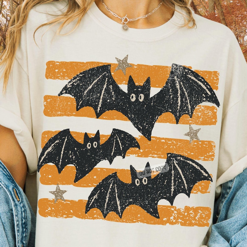 May include: A cream-colored t-shirt featuring a graphic print of three black bats flying across horizontal, distressed orange stripes. The design includes small, scattered gray stars and a subtle, semi-transparent watermark text overlay.