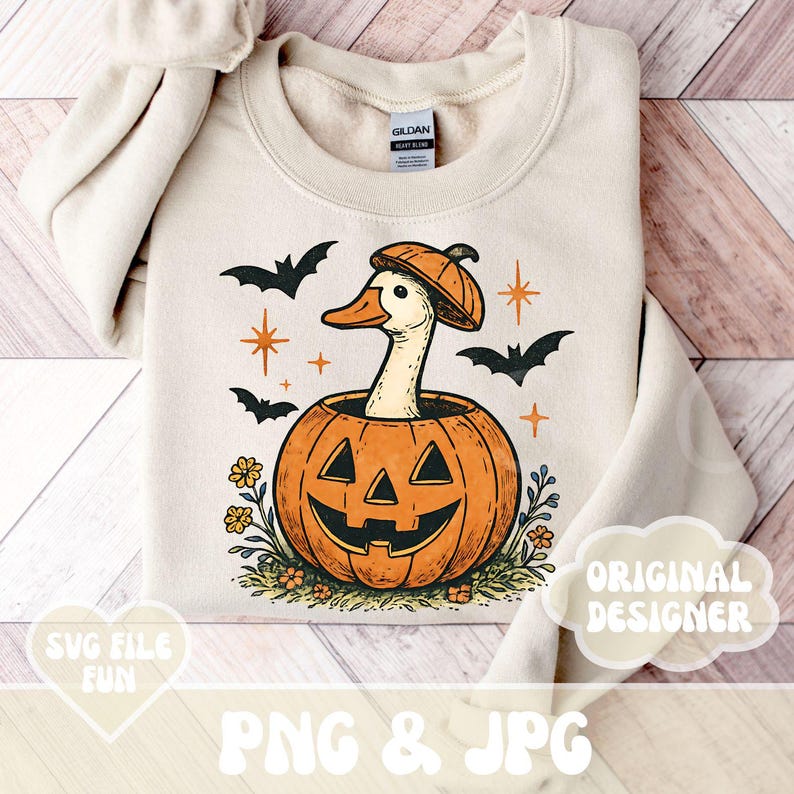 May include: Cream-colored sweatshirt featuring a Halloween-themed graphic. The design includes a duck wearing a pumpkin hat, emerging from a carved pumpkin. Black bats and orange stars surround the central image. The words "PNG & JPG" and "SVG FILE FUN" are also visible.