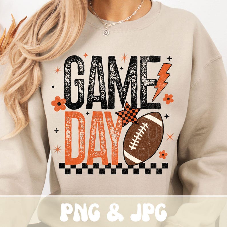 May include: Beige sweatshirt with the words "GAME DAY" in black and orange, a brown football, and orange lightning bolt and flower graphics. The design includes a checkered star and a black and orange checkered border.