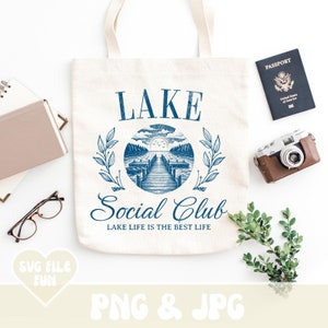 May include: A white tote bag with a blue graphic design that says "LAKE Social Club LAKE LIFE IS THE BEST LIFE". The graphic includes a lake scene with a dock and a sunset.