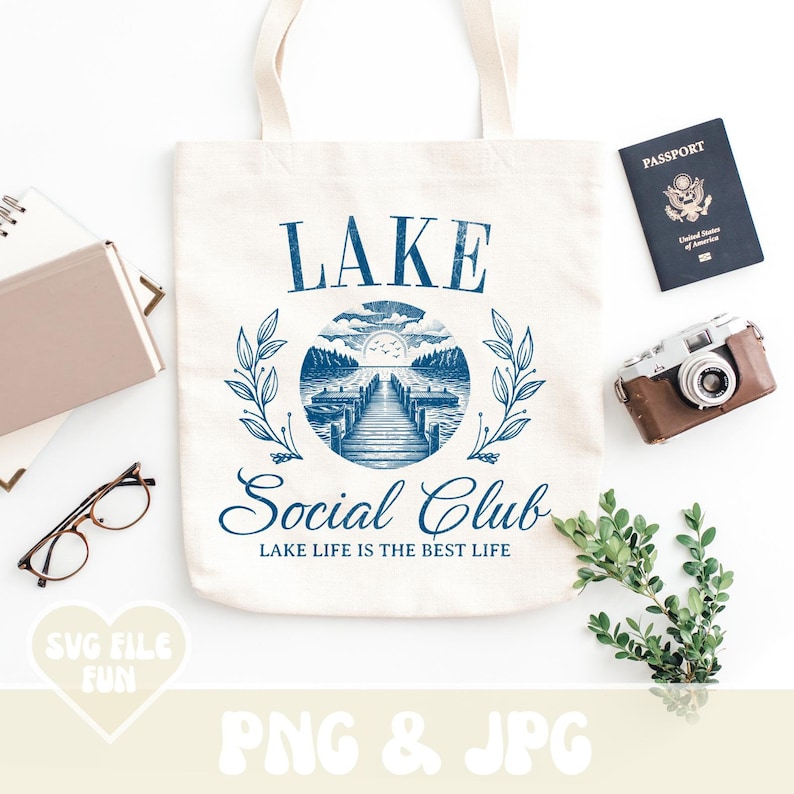 May include: A white tote bag with a blue graphic design that says "LAKE Social Club LAKE LIFE IS THE BEST LIFE". The graphic includes a lake scene with a dock and a sunset.