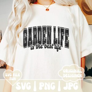 May include: A white t-shirt with black text that reads "GARDEN LIFE is the best life". The text has a distressed, vintage look. The shirt is being worn by a person with long blonde hair.