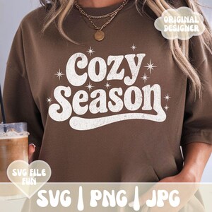 Puede incluir: Una sudadera marr&oacute;n con la frase "Cozy Season" en blanco, con una fuente retro. La imagen tambi&eacute;n incluye el texto "SVG | PNG | JPG" y las palabras "SVG FILE FUN" en un gr&aacute;fico en forma de coraz&oacute;n.