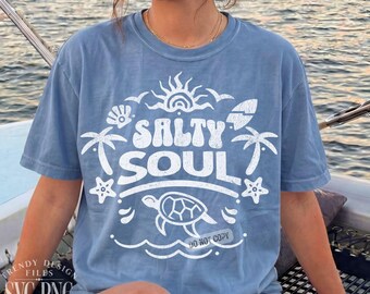 May include: A pale blue t-shirt featuring the words "Salty Soul" in white, accompanied by illustrations of a sun, palm trees, a seashell, a surfboard, a starfish, a turtle, and waves. The shirt has a loose fit.