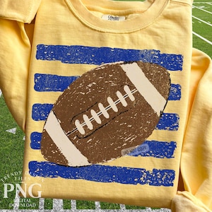 K&ouml;nnte beinhalten: Ein blassgelbes Langarm-Sweatshirt mit einer braunen American-Football-Grafik, die &uuml;ber vier horizontalen, abgenutzten blauen Streifen zentriert ist. Der Football hat wei&szlig;e Schn&uuml;rsenkel und wei&szlig;e Endstreifen. Das Innenetikett zeigt Comfort Colors.