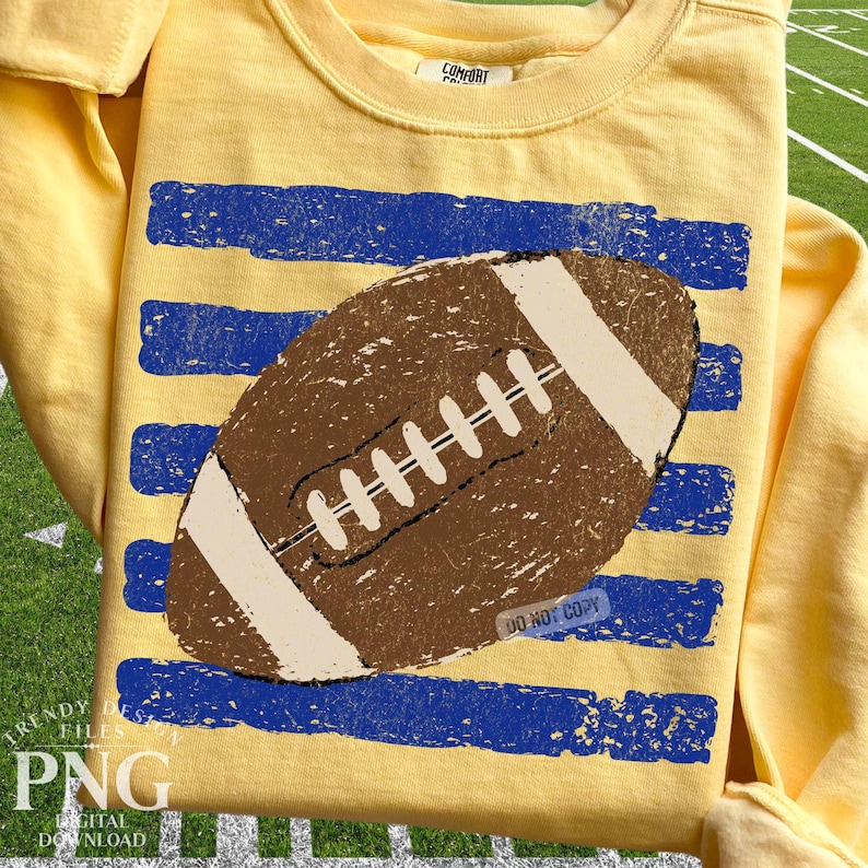 K&ouml;nnte beinhalten: Ein blassgelbes Langarm-Sweatshirt mit einer braunen American-Football-Grafik, die &uuml;ber vier horizontalen, abgenutzten blauen Streifen zentriert ist. Der Football hat wei&szlig;e Schn&uuml;rsenkel und wei&szlig;e Endstreifen. Das Innenetikett zeigt Comfort Colors.