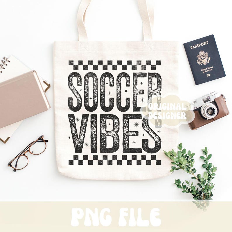 K&ouml;nnte beinhalten: Eine cremefarbene Tragetasche mit dem Text "SOCCER VIBES" in einer verwaschenen schwarzen Schrift. Die Tasche wird von einem Reisepass, einer Vintage-Kamera, einer Brille, einem Notizbuch und Gr&uuml;nzeug auf einer wei&szlig;en Oberfl&auml;che begleitet.