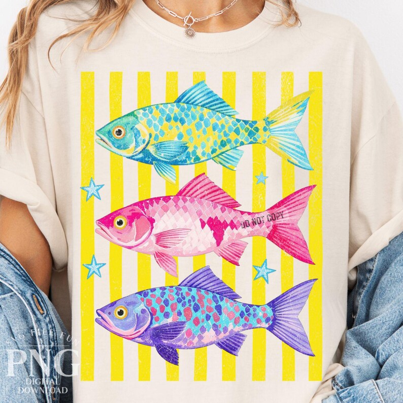 May include: Cream-colored t-shirt featuring three colorful fish: turquoise, pink, and purple, set against yellow vertical stripes. The fish have detailed scales and fins. Blue stars are scattered around the fish. The text "DO NOT COPY" is on the pink fish.