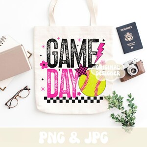 May include: A white canvas tote bag with the words "GAME DAY" in black and pink, featuring a softball graphic. The bag is decorated with pink flowers, a lightning bolt, and a checkered star. A passport, camera, and greenery are next to the bag.