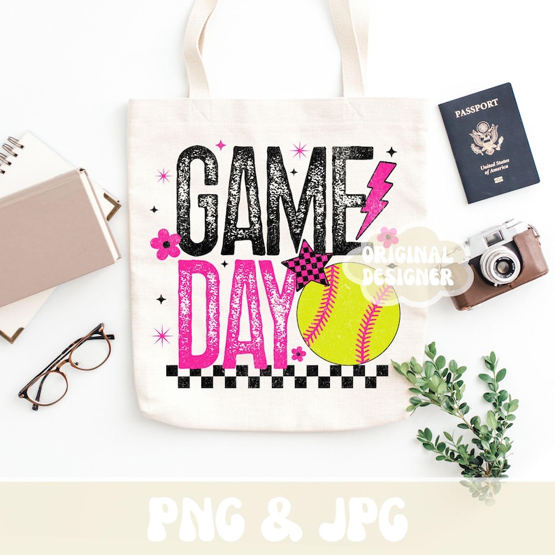 May include: A white canvas tote bag with the words "GAME DAY" in black and pink, featuring a softball graphic. The bag is decorated with pink flowers, a lightning bolt, and a checkered star. A passport, camera, and greenery are next to the bag.