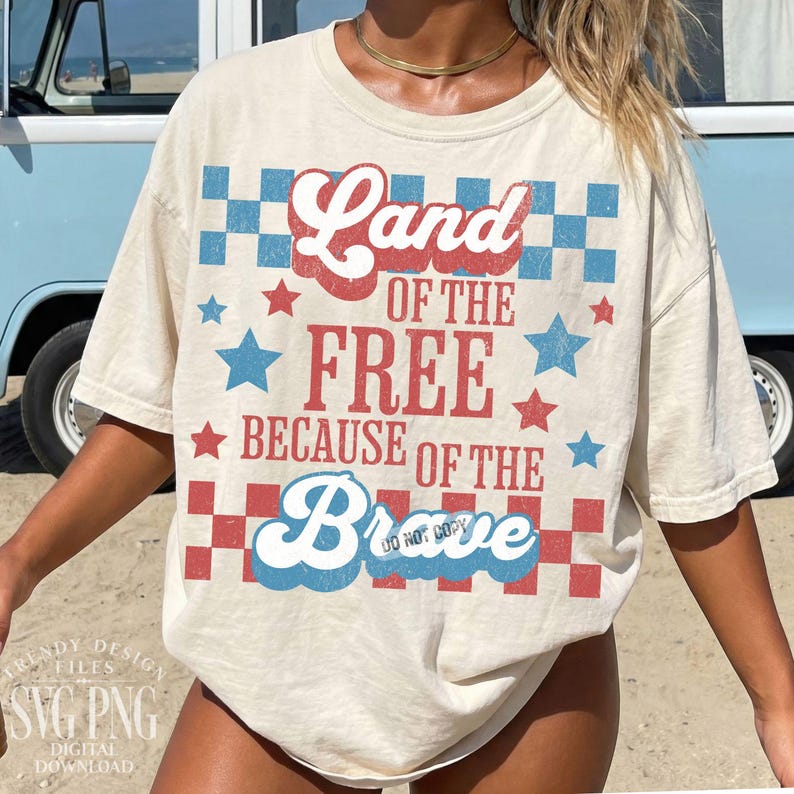 May include: Cream-colored t-shirt with the text "Land of the Free Because of the Brave" in red and blue. The design includes stars and checkered patterns in red and blue, evoking a patriotic theme. The shirt has a vintage, slightly faded look.