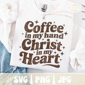 May include: White sweatshirt with the text "Coffee in my hand, Christ in my heart" in brown, surrounded by starbursts. The sweatshirt has a crew neck and long sleeves. The bottom of the image shows the file types: SVG, PNG, and JPG.