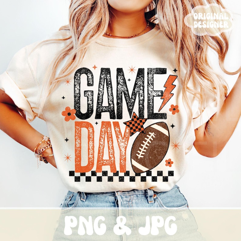 May include: Cream-colored t-shirt with the words "GAME DAY" in black and orange, featuring a football graphic and a lightning bolt. The shirt has a vintage, distressed design with floral and star accents.