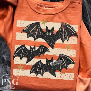 May include: An orange crewneck sweatshirt featuring a graphic print of three black bats with textured wings. The design includes horizontal off-white distressed stripes and small gold stars scattered across the front of the garment.
