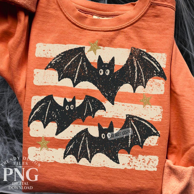 May include: An orange crewneck sweatshirt featuring a graphic print of three black bats with textured wings. The design includes horizontal off-white distressed stripes and small gold stars scattered across the front of the garment.