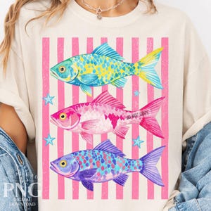 May include: Cream-colored t-shirt featuring three colorful fish designs. The fish are in shades of blue, pink, and purple, with yellow and white accents. The background has pink and white vertical stripes and small blue star shapes.