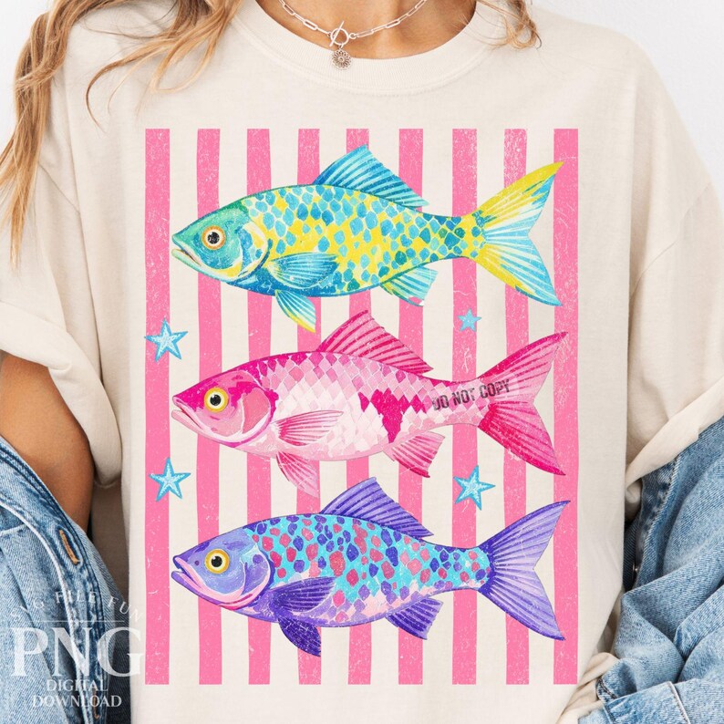 May include: Cream-colored t-shirt featuring three colorful fish designs. The fish are in shades of blue, pink, and purple, with yellow and white accents. The background has pink and white vertical stripes and small blue star shapes.