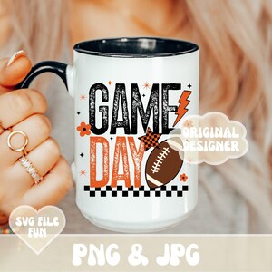 May include: White ceramic mug with a black handle and rim, featuring the words "GAME DAY" in black and orange, along with a football graphic and lightning bolt. The mug has a football theme.