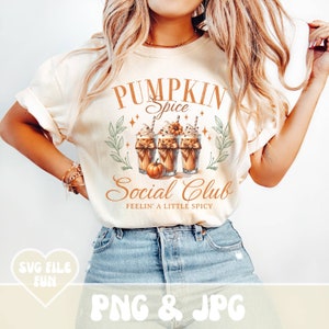 May include: Cream-colored t-shirt featuring a graphic with the text "Pumpkin Spice Social Club" and images of pumpkin spice lattes. The design includes decorative elements like stars and leaves, with the phrase "Feelin' a Little Spicy" below.