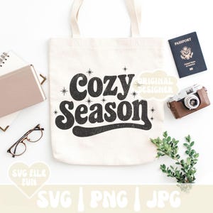 Puede incluir: Un bolso tote de color crema con las palabras "Cozy Season" en una fuente negra retro. La bolsa est&aacute; rodeada por un pasaporte, una c&aacute;mara vintage, gafas, un cuaderno y vegetaci&oacute;n. Tambi&eacute;n se ven las palabras "SVG FILE FUN" y "SVG | PNG | JPG".