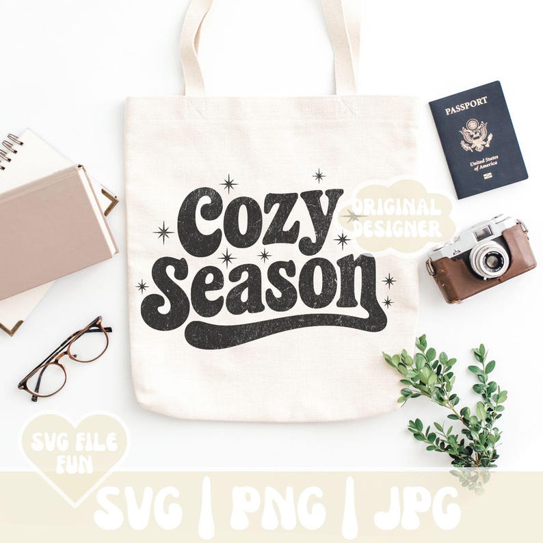 Puede incluir: Un bolso tote de color crema con las palabras "Cozy Season" en una fuente negra retro. La bolsa est&aacute; rodeada por un pasaporte, una c&aacute;mara vintage, gafas, un cuaderno y vegetaci&oacute;n. Tambi&eacute;n se ven las palabras "SVG FILE FUN" y "SVG | PNG | JPG".