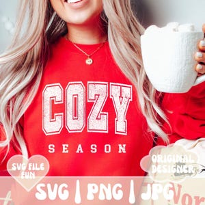 May include: A red sweatshirt with the text "Cozy Season" in white distressed lettering. The sweatshirt is being worn by a person holding a white mug with marshmallows. The image also includes the text "SVG | PNG | JPG" and "Original Designer".