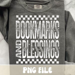 以下が含まれることがあります： 「BOOKMARKS AND BLESSINGS」の文字が白の掠れたフォントでプリントされたダークグレーのスウェットシャツ。テキストは白い市松模様で縁取られています。スウェットシャツの下部には「PNG FILE」と書かれています。