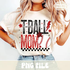 May include: Cream-colored t-shirt with the text "T-BALL MOM" in black and red. A baseball graphic is featured, along with a red lightning bolt and floral accents. The shirt has rolled-up sleeves.