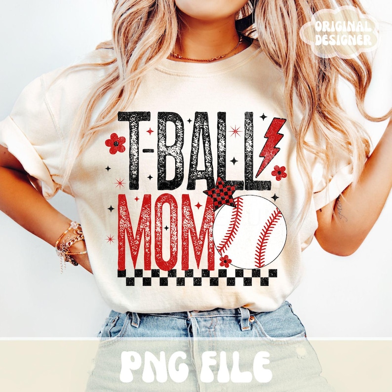 May include: Cream-colored t-shirt with the text "T-BALL MOM" in black and red. A baseball graphic is featured, along with a red lightning bolt and floral accents. The shirt has rolled-up sleeves.