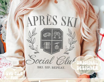 Könnte beinhalten: Ein weißer Pullover mit einem grauen Grafikdesign, auf dem "Aprés Ski Social Club" steht, mit einem Wappen, das Skistöcke, eine Skibrille, Weingläser und einen Skilift zeigt. Der Text unter dem Wappen lautet "Ski. Sip. Repeat."