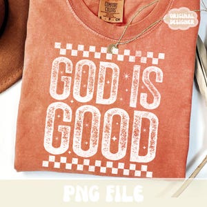 K&ouml;nnte beinhalten: Ein korallenfarbenes T-Shirt mit dem Schriftzug "GOD IS GOOD" in gro&szlig;en, verwaschenen wei&szlig;en Buchstaben. Das Design umfasst ein Schachbrettmuster und Sternakzente. Eine goldene Halskette und ein Etikett mit der Aufschrift "ORIGINAL DESIGNER" sind ebenfalls sichtbar.