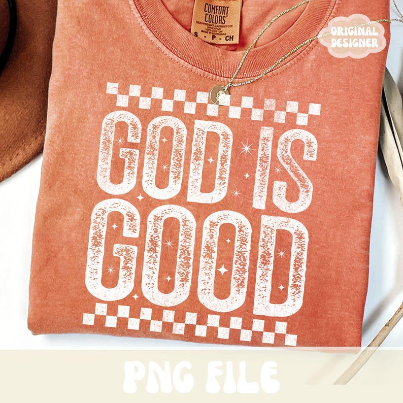 K&ouml;nnte beinhalten: Ein korallenfarbenes T-Shirt mit dem Schriftzug "GOD IS GOOD" in gro&szlig;en, verwaschenen wei&szlig;en Buchstaben. Das Design umfasst ein Schachbrettmuster und Sternakzente. Eine goldene Halskette und ein Etikett mit der Aufschrift "ORIGINAL DESIGNER" sind ebenfalls sichtbar.