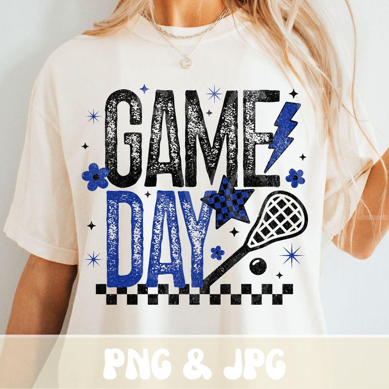 May include: Cream-colored t-shirt with the words "GAME DAY" in black and blue, featuring a lacrosse stick, star, and floral accents. The design includes a checkered pattern and lightning bolt graphic.