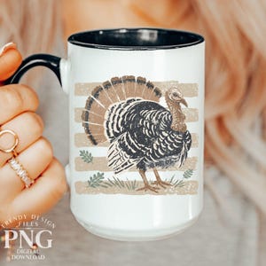May include: A white ceramic mug with a black handle and interior rim. The exterior features a graphic of a turkey in shades of brown and black, set against a background of horizontal tan stripes and small green leaf accents.