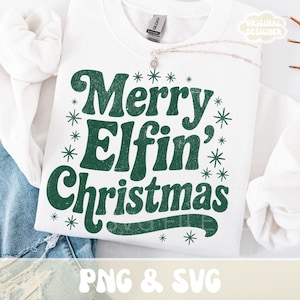 May include: White sweatshirt with the text "Merry Elfin' Christmas" in green, surrounded by snowflakes. A silver necklace with a small pendant is visible. The sweatshirt is folded over denim jeans.