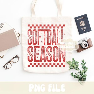May include: A white canvas tote bag with the words "SOFTBALL SEASON" in red, distressed lettering. The bag is surrounded by a passport, a vintage camera, a notebook, glasses, and greenery.