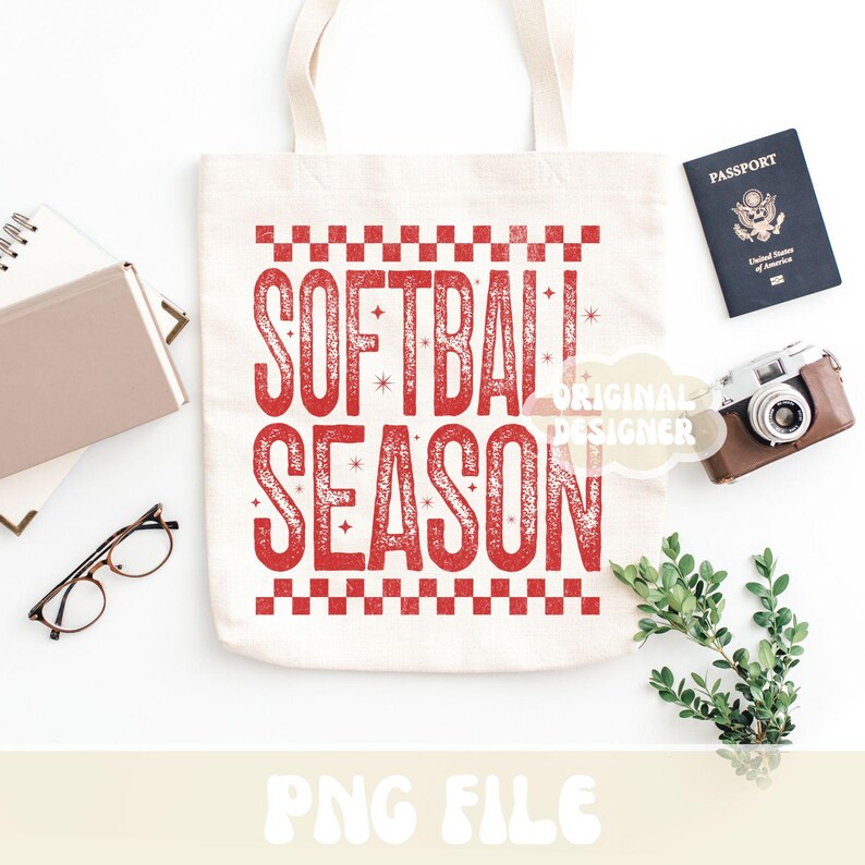 May include: A white canvas tote bag with the words "SOFTBALL SEASON" in red, distressed lettering. The bag is surrounded by a passport, a vintage camera, a notebook, glasses, and greenery.