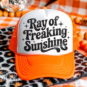 May include: Orange and white trucker hat with the text "Ray of Freaking Sunshine" in black. The hat has a mesh back and a solid orange brim. The text is in a bold, retro font with star accents.