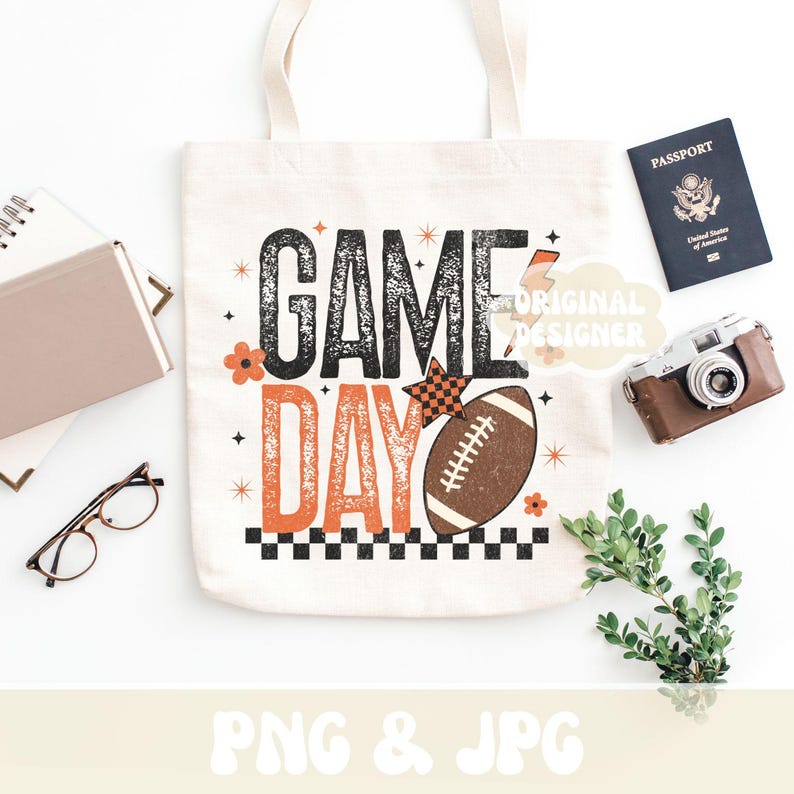 May include: A white tote bag with the words "GAME DAY" in black and orange, featuring a football graphic. The bag is surrounded by a passport, a vintage camera, and other accessories.