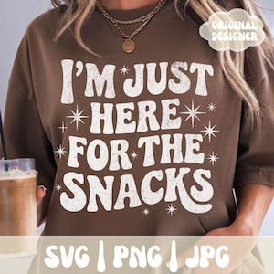 May include: Brown sweatshirt with the text "I'M JUST HERE FOR THE SNACKS" in white, retro-style lettering. The design includes starburst accents. The words "SVG | PNG | JPG" are at the bottom. A drink is held in the left side of the image.