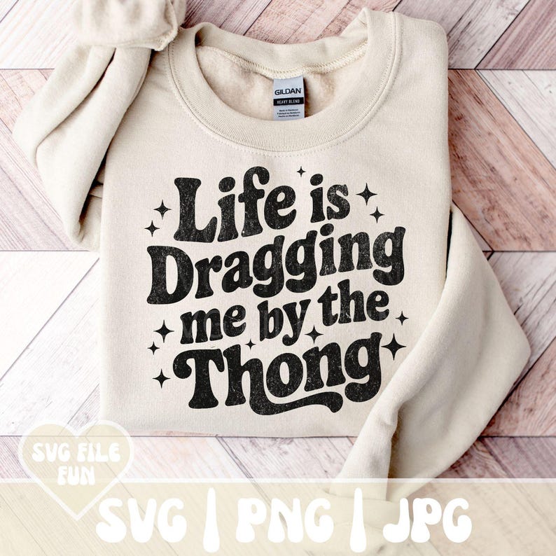 May include: A cream-colored sweatshirt with the phrase "Life is Dragging me by the Thong" in black, retro-style lettering. The design includes star accents. The sweatshirt is folded on a wooden surface.