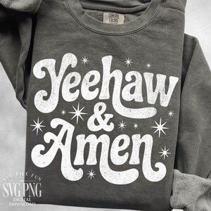 Puede incluir: Una sudadera gris oscuro con las palabras "Yeehaw & Amen" en blanco, con una fuente retro. El dise&ntilde;o incluye gr&aacute;ficos de estrellas. La sudadera est&aacute; hecha de un material suave y tiene cuello redondo.
