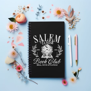 K&ouml;nnte beinhalten: Schwarzer Spiralblock mit einem wei&szlig;en Design, auf dem "Salem Witch Book Club Est. 1692 Bell, Book & Candle" steht. Das Design zeigt ein Buch mit einem Mond und Sternen dar&uuml;ber. Der Block ist von Blumen und Stiften auf hellblauem Hintergrund umgeben.