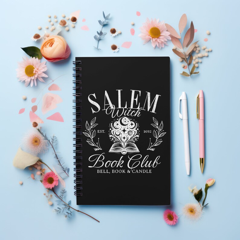 K&ouml;nnte beinhalten: Schwarzer Spiralblock mit einem wei&szlig;en Design, auf dem "Salem Witch Book Club Est. 1692 Bell, Book & Candle" steht. Das Design zeigt ein Buch mit einem Mond und Sternen dar&uuml;ber. Der Block ist von Blumen und Stiften auf hellblauem Hintergrund umgeben.